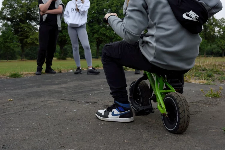 Teenager on undersized bike with two others looking on, all on overgrown asphalt [photograph]