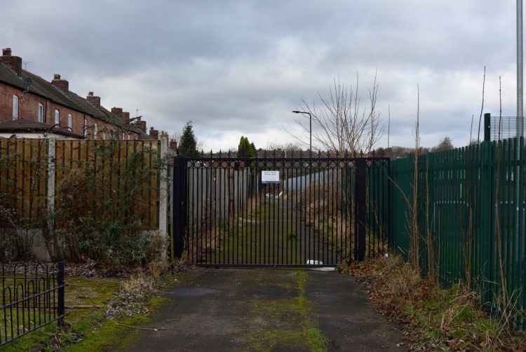 A black gate to an overgrown alleyway running behind terraced housing in South Yorkshire