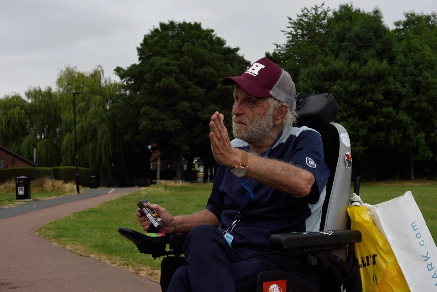 Older man in cap, holding cigarettes and gesturing from his electric wheelchair with shopping bags attached [photograph]