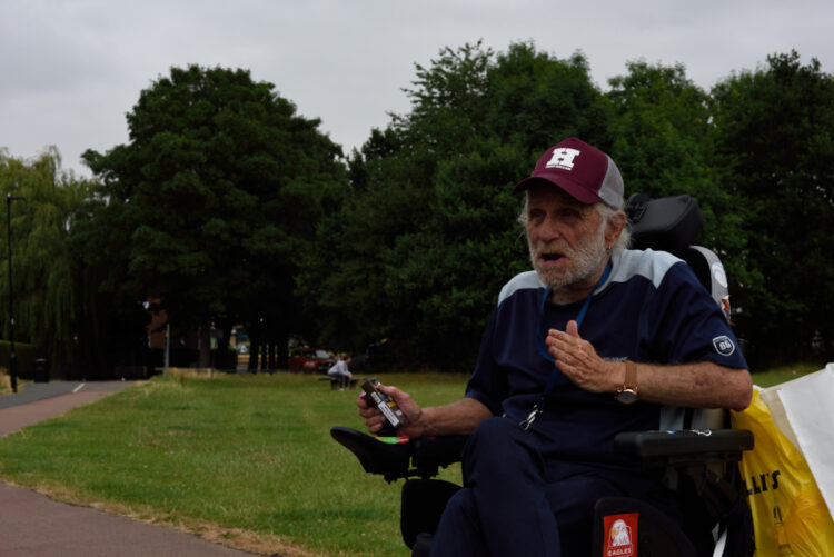 man in electric wheelchair wearing a baseball cap talks and gestures to somebody listening out of shot