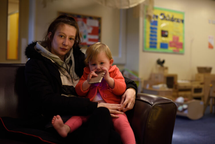 mother and toddler daughter sit on a sofa looking to camera while the toddler chews a mobile phone case