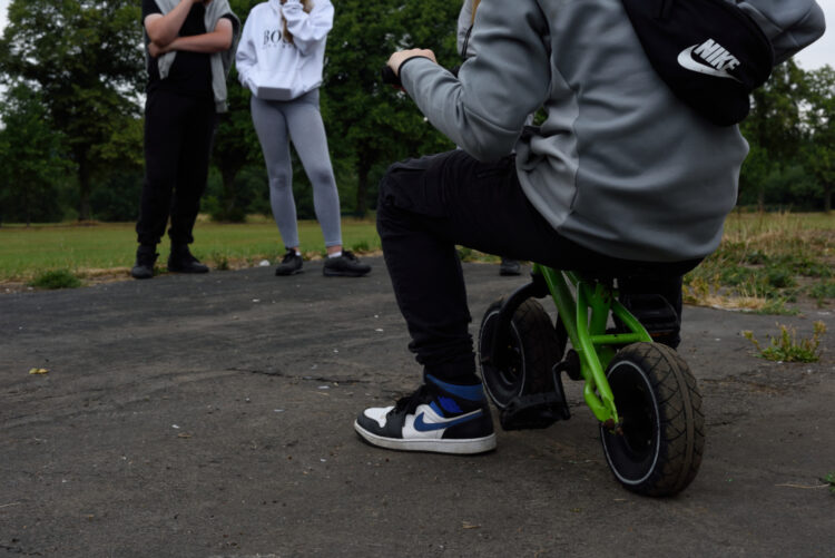 Teenagers in a park. A boy on a bike that is too small for him faces two other young people. They are all standing on a patch of concrete