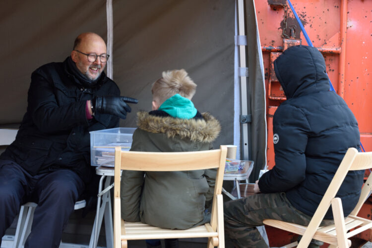 Man laughs and gestures as he talks to two young boys under cover of a gazebo. They all wear thick goats