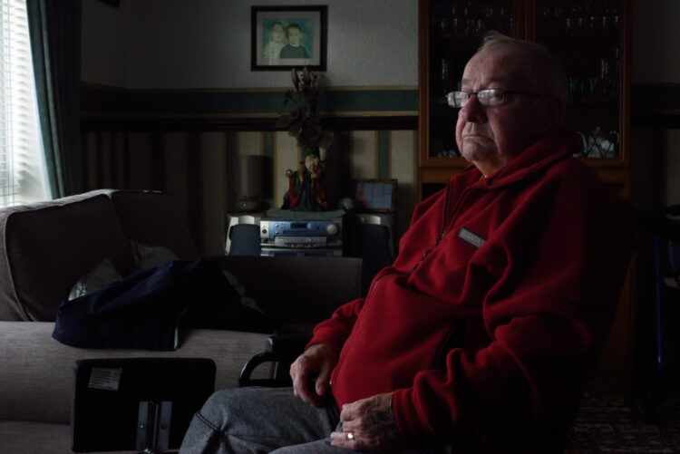 older man sits alone looking off to side out of his window