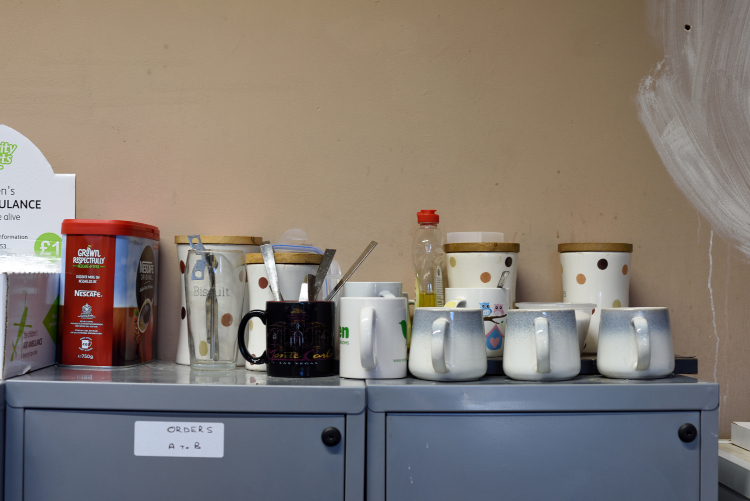 Selection of mismatched mugs and items for making hot drinks on top of a pair of filing cabinets