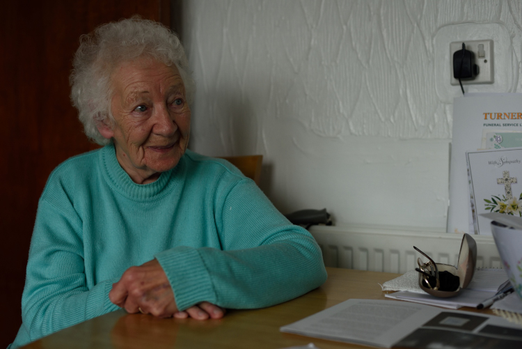 older woman sitting at kitchen table looking at somebody opposite her who is out of shot