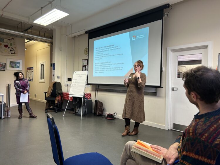 White woman stands before a screen with a Who is Your Neighbour presentation on it, addressing a seated group of people on a training day