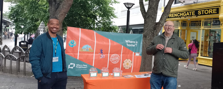 A black man and a white man stand smiling on either side of a stall in a shopping precinct. The stall is a bright orange table with a banner behind asking the question Where Is Home?